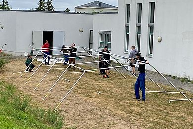 8 Personen die ein Zeltgestänge, hinter einem Haus aufbauen