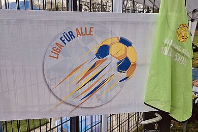Logo Liga für Alle hängt am Zaum vor dem Kunstrasenplatz