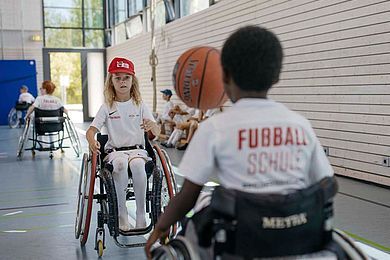 Zwei Teilnehmende sitzen im Rollstuhl und probieren Rollstuhl-Basketball aus