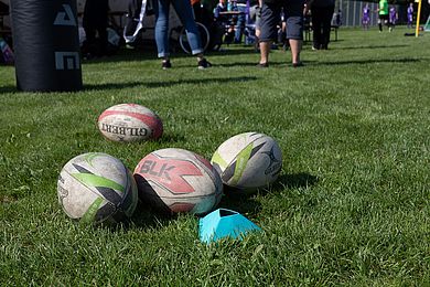 Auf einer Wiese liegen verschieden farbige Rugbybälle. Im Hintergrund sieht man die Füße und Beine vom Besucher*innen.
