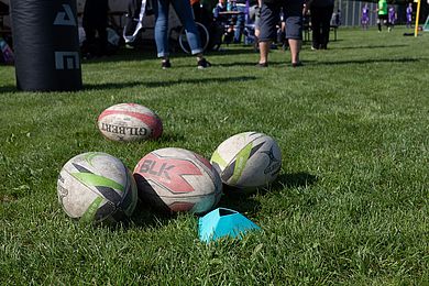 Auf einer Wiese liegen verschieden farbige Rugbybälle. Im Hintergrund sieht man die Füße und Beine vom Besucher*innen.