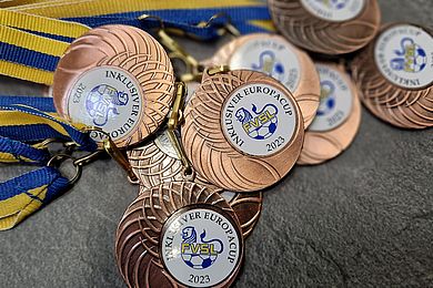 Bronzefarbene Medaillen mit der Aufschrift "Inklusiver Europacup 2023", in der Mitte das FVSL Logo. Die Bänder der Medaillen sind blau und gelb.