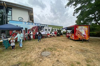 Vor der Sporthalle stehen verschiedene Tische für die Speiseversorgung und ein roter Eiswagen