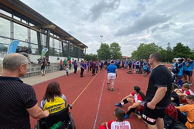 Menschen mit und ohne Behinderung sind auf einem Sportplatz, vor einer Sporthalle versammelt