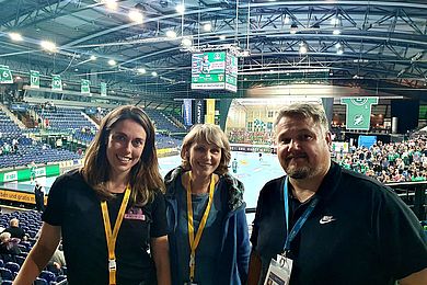 2 Frauen und ein Mann stehen im Publikum in einer großen Sporthalle