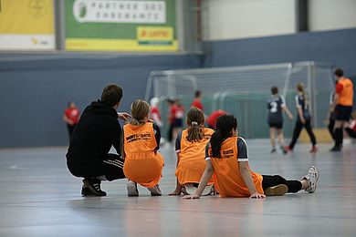 Drei Kinder und ein Erwachsener sitzen und hocken auf dem Fußboden der Sporthalle und beobachten andere Spieler*innen