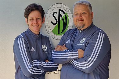 Links steht Franka Schmidt, rechts daneben Axel Ackermann. Sie tragen grauen Sportanzüge