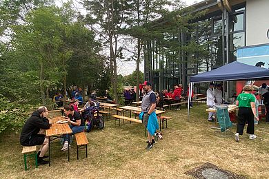 viele Bierzeltgarnituren stehen unter Bäumen. Rechts steht ein blauer Pavillon, unter dem Kuchen verkauft wird