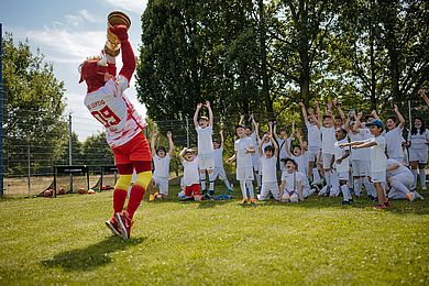 RB Leipzig-Maskottchen Bulli hält einen Pokal in die Luft, im Hintergrund jubeln die Kinder