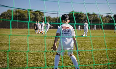 Ein Junge im Tor. Im Hintergrund nimmt ein Teilnehmer Anlauf, um den Ball zu schießen.