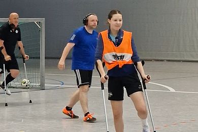 ein Mädchen spielt mit Unterarmstützen Fußball. Ein Mann läuft hinter ihr entlang