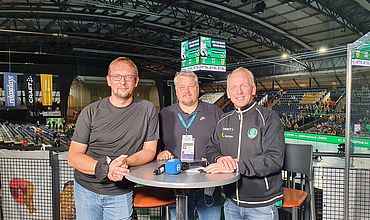 drei Männer stehen an einem Stehtisch in einer großem Halle mit einem Mikrofon auf dem Tisch. Alle tragen dunkelgraue Oberteile und blaue Jeans